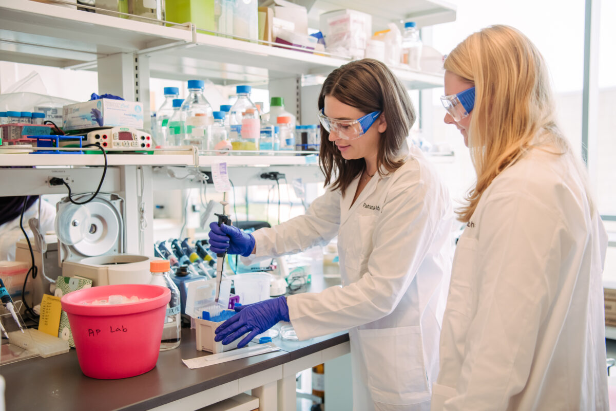 Two women at a lab bench; one operates a pipette while the other looks on.