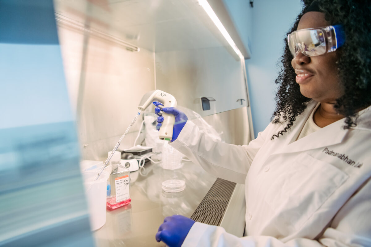 Woman in lab coat and purple gloves reaches under the partial glass wall of a fume hood holding a pipette.