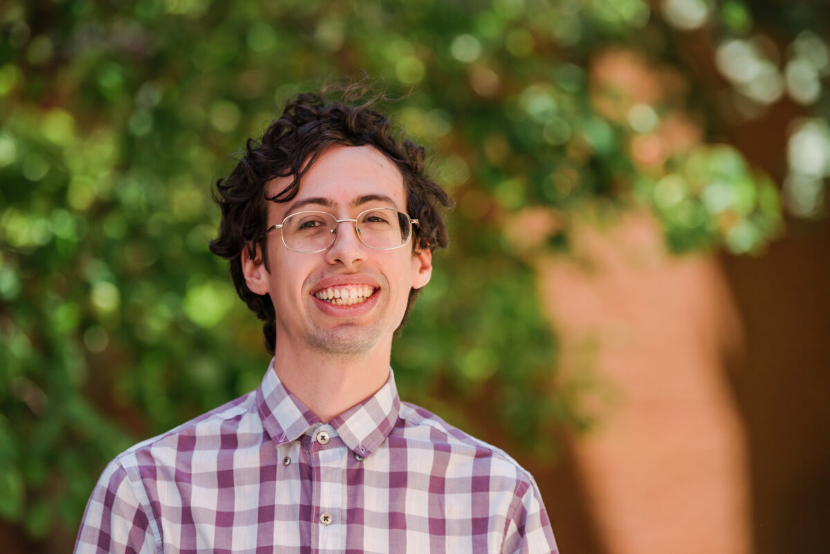 A young man wearing light colored glasses and a purple and white checkered dress shirt stands in front of a brick building and some trees.