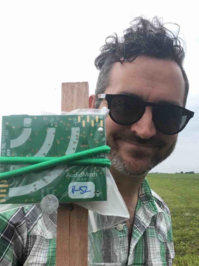 Man in sunglasses standing next to a small green circuit board attached to a wooden post in the foreground