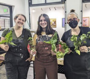 group photo of three women in overalls holding plants, indoors