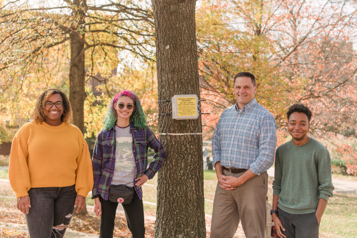 group photo of four people. A tree trunk is in the center with a small silver box attached to it and a metal ribbon wrapped around it. Fall foliage in the background.