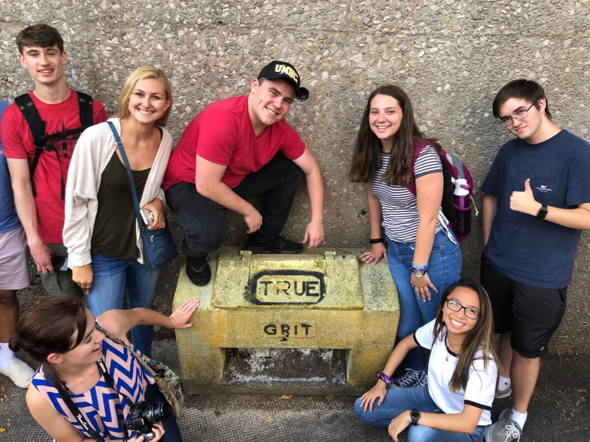 A group of seven college students gather around a yellow cement box with the words True Grit written in black paint.