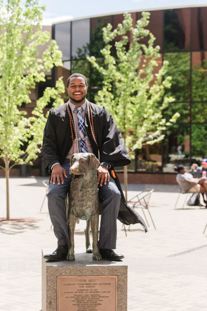 an alum in a graduation gown sits on top of a bronze statue of a dog