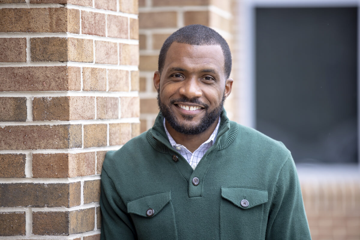 A man in a green button down shirt leans against a brick wall