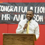 a man in a white dress shirt and a yellow tie stands at a lectern with a sign behind him that reads Congratulations Mr. Anderson. Derek Anderson was selected as Principal of the year for Maryland.