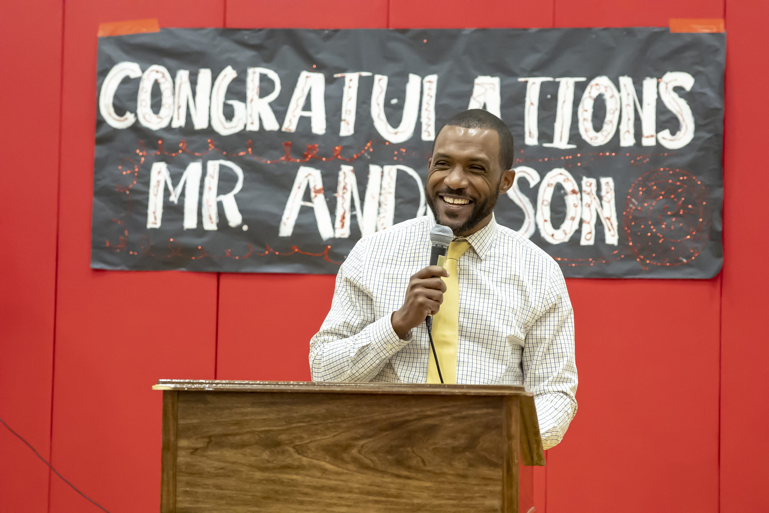 a man in a white dress shirt and a yellow tie stands at a lectern with a sign behind him that reads Congratulations Mr. Anderson. Derek Anderson was selected as Principal of the year for Maryland.