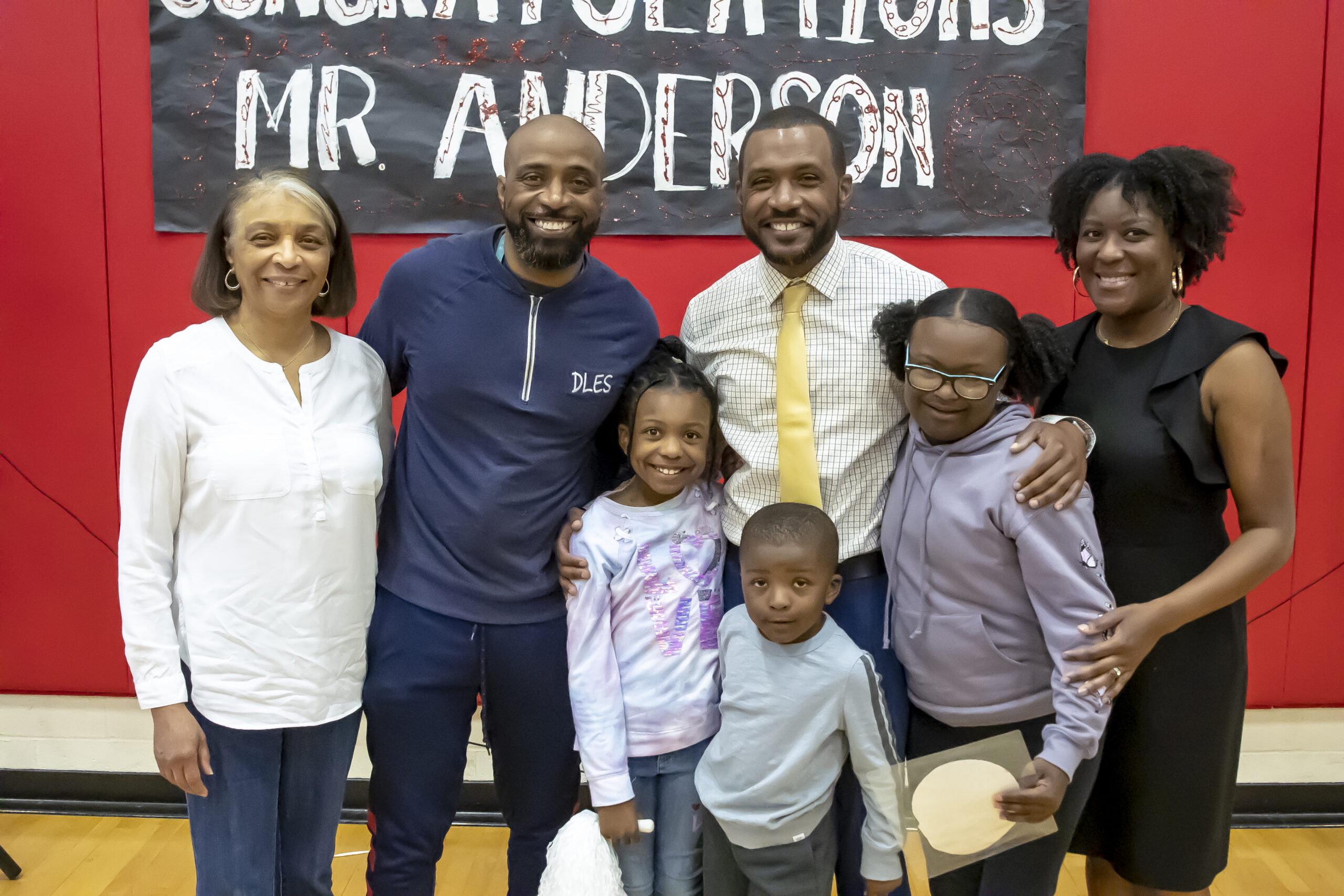 a man in a white dress shirt and a yellow tie stands with his family and a sign behind them that reads Congratulations Mr. Anderson