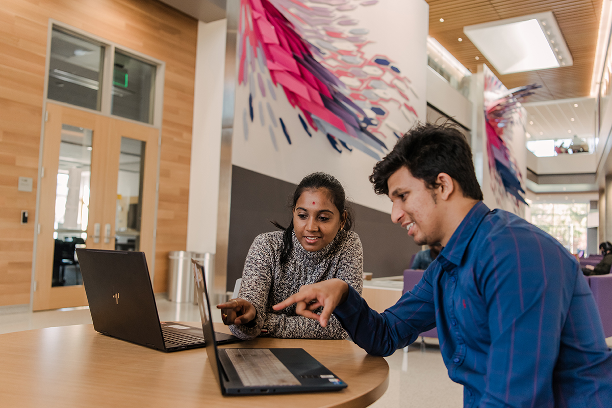 A young woman and young man sitting together, pointing at their laptops.