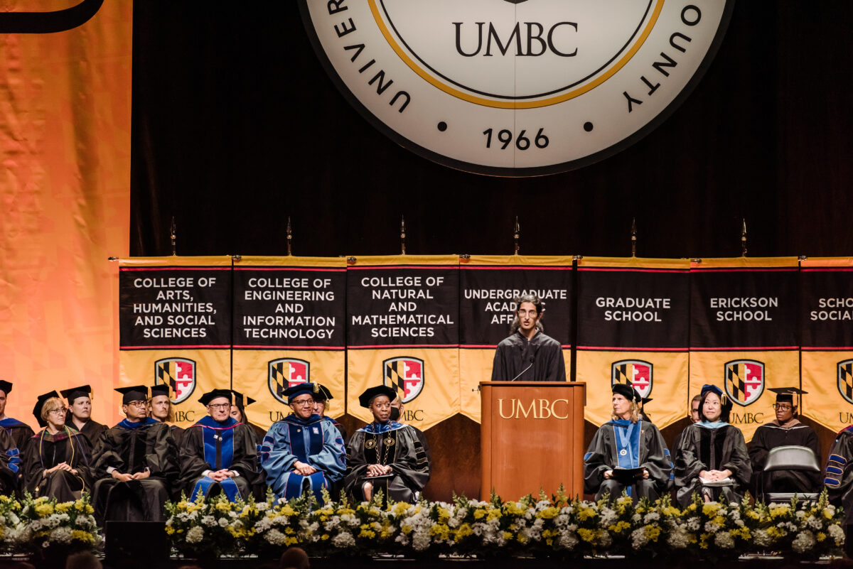 Tall student stands at a UMBC podium wearing a robe with university officials sitting behind them and UMBC seal and flags in the background