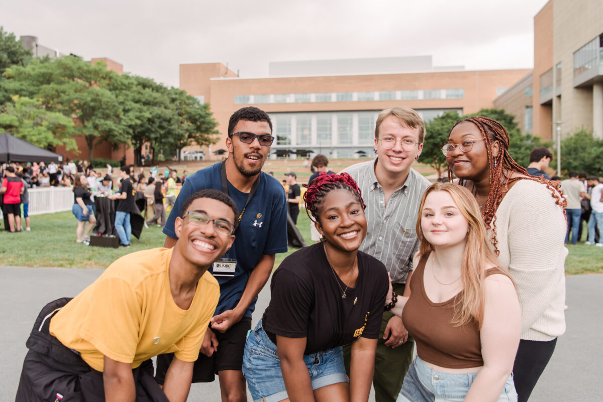 Six students pose for a smiling group photo outdoors in front of a campus building