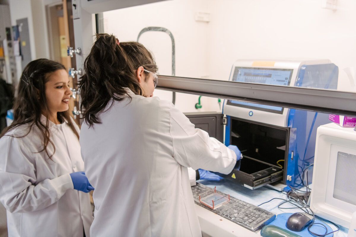 two women in lab coats work at a large instrument inside a hood.