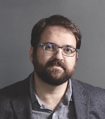 A man with dark hair, a beard and glasses looks at the camera