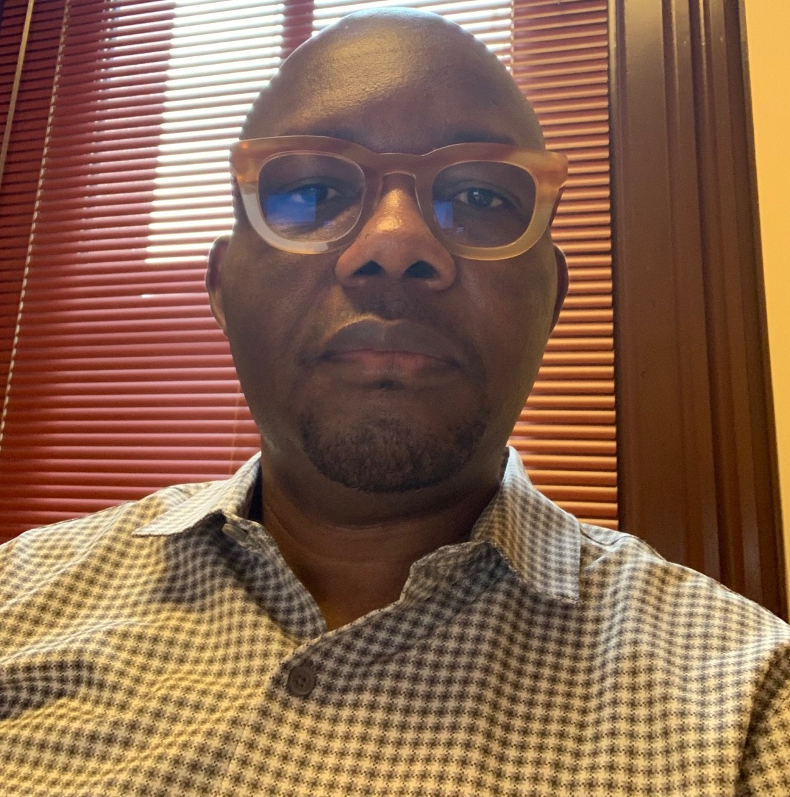 A Black man with glasses and a checkered shirt looks at the camera