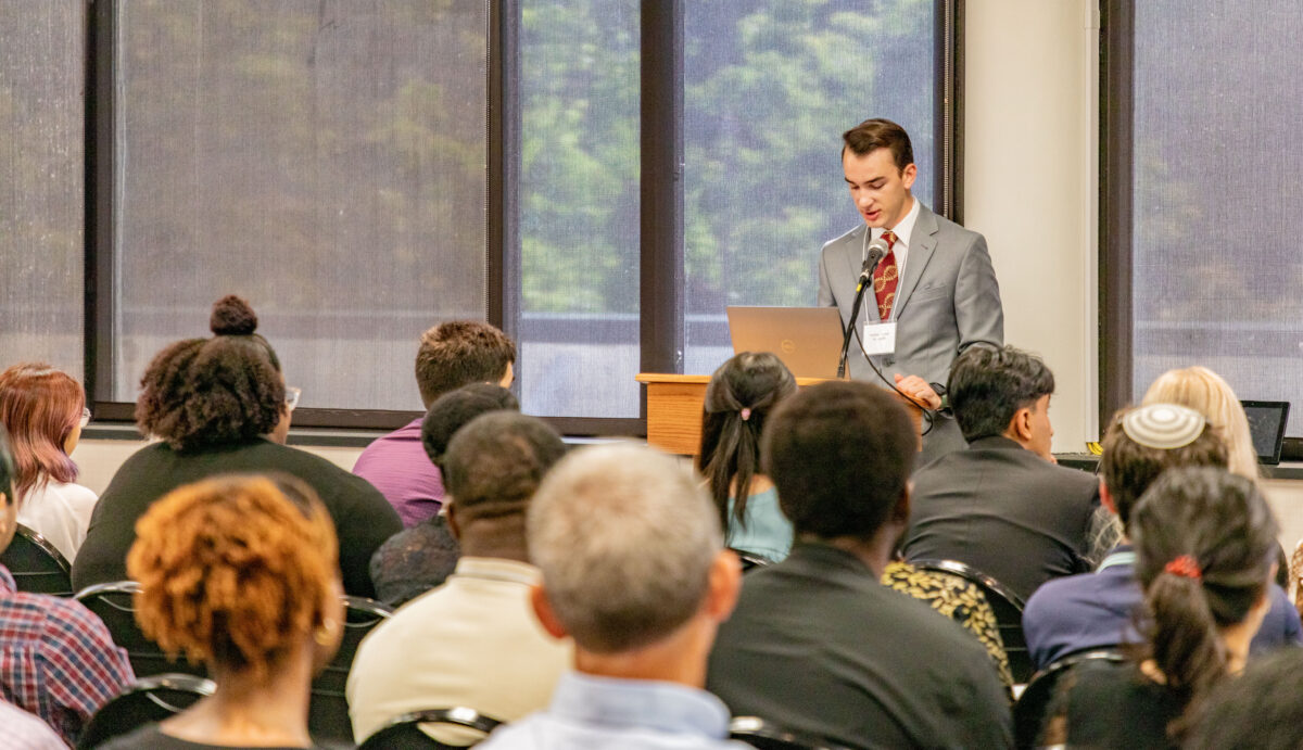 young man at podium speaking into microphone; rows of seated people in front of him 