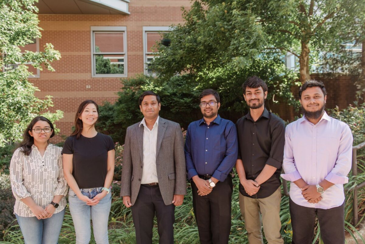 A group of 6 people stand in a line and smile at the camera.