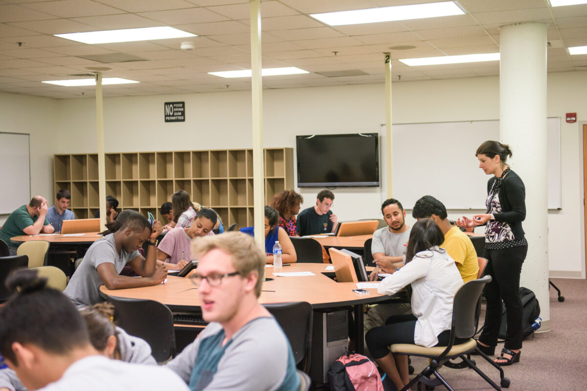 groups of students seated at tables engaged in discussion; an instructor stands at one table, talking with students