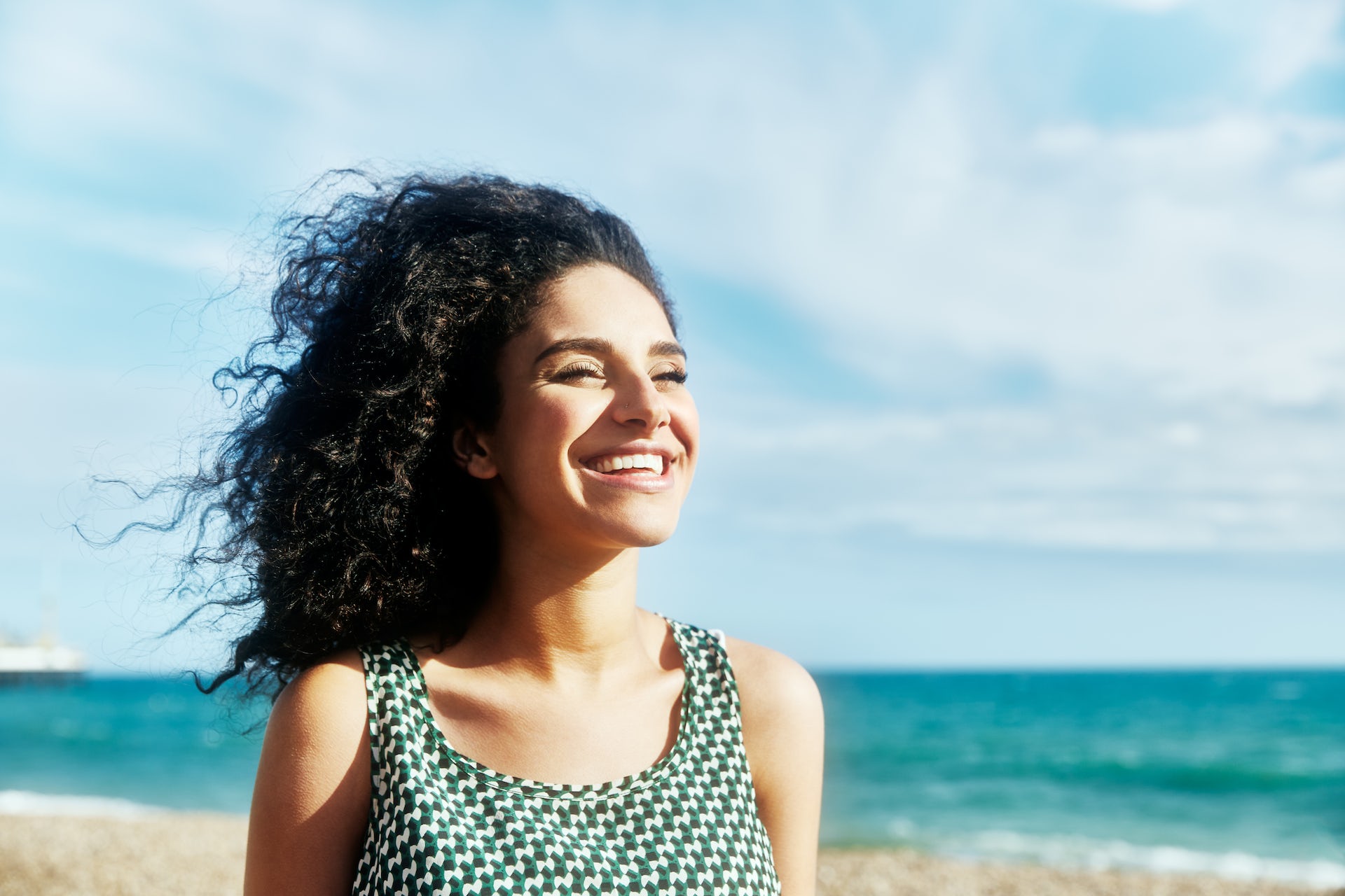 smiling woman standing on a beach, her curly hair blowing in the wind