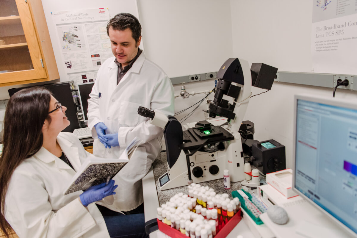 Two people in lab coats and gloves in a laboratory; a microscope, vials containing flies, and  a computer sit on the counter.