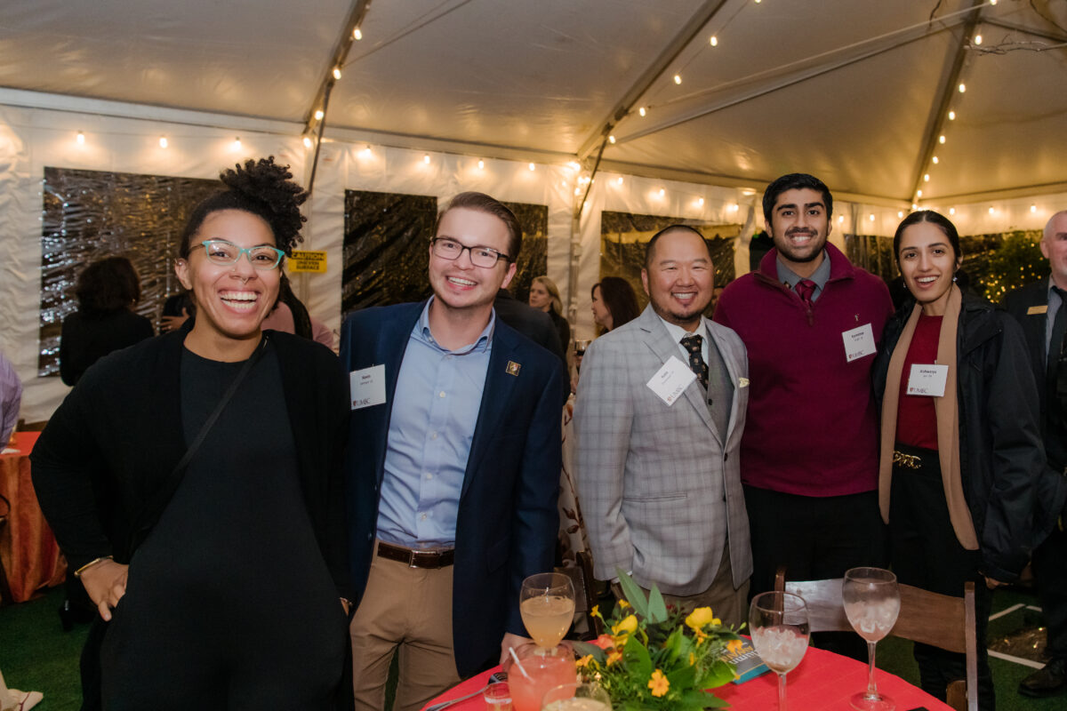 young professionals smile in a outdoor event tent with fancy lights strung up behind them.