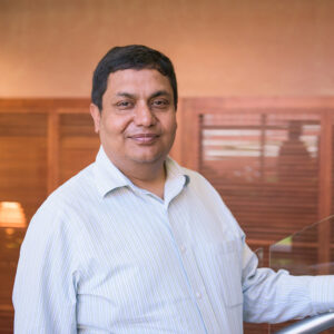Profile portrait of Anupam Joshi, standing in front of a reflective tan wall.