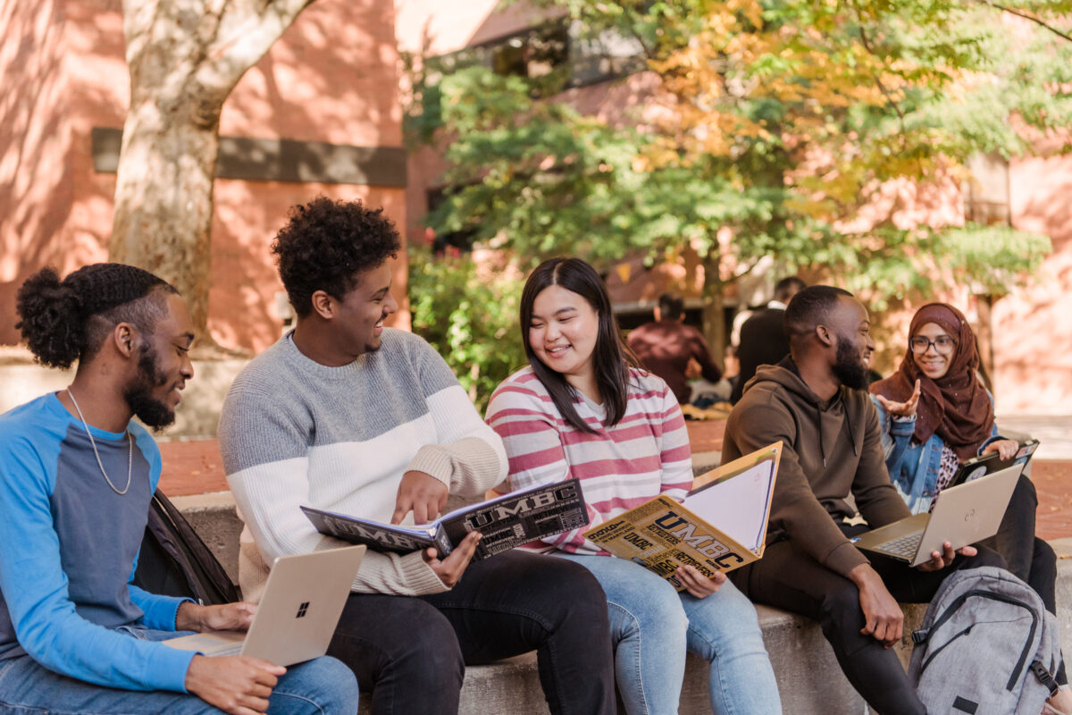 Five UMBC students sitting amongst one another on UMBC's campus. Students have books and laptops in their hands with UMBC's logo on several books.