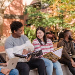 Five UMBC students sitting amongst one another on UMBC's campus. Students have books and laptops in their hands with UMBC's logo on several books.