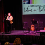 Woman wearing a mask while standing on stage behind a podium in front of a screen. The screen says "moving stories: latinas en Baltimore"