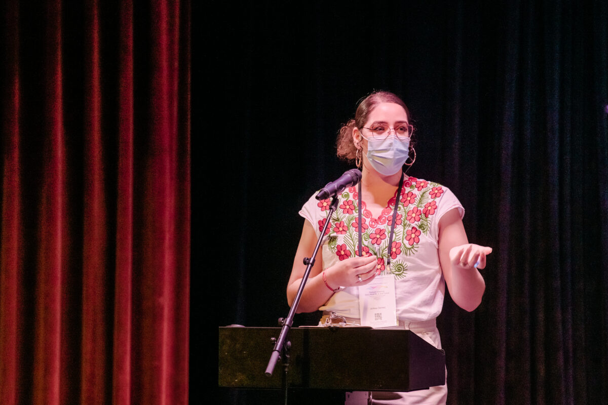 Woman wearing glasses and a masking, standing on a stage behind a podium and mic. 