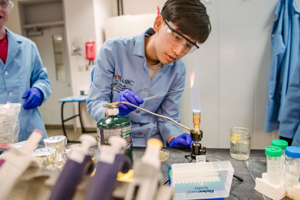 A student in a lab coat holds a glass rod in the flames of a Bunsen burner