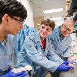 Three people in lab coats examine equipment at a lab bench.