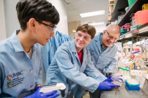 Three people in lab coats examine equipment at a lab bench.