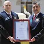 Two mean wearing formal attire hold a framed certificate.