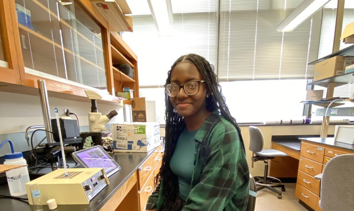 woman in green sits at a lab bench, looking at the camera--she is a Roth award recipient for summer research.
