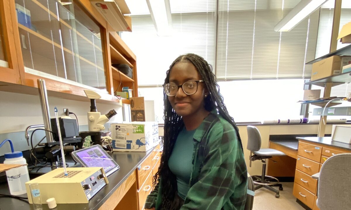 woman in green sits at a lab bench, looking at the camera--she is a Roth award recipient for summer research.
    