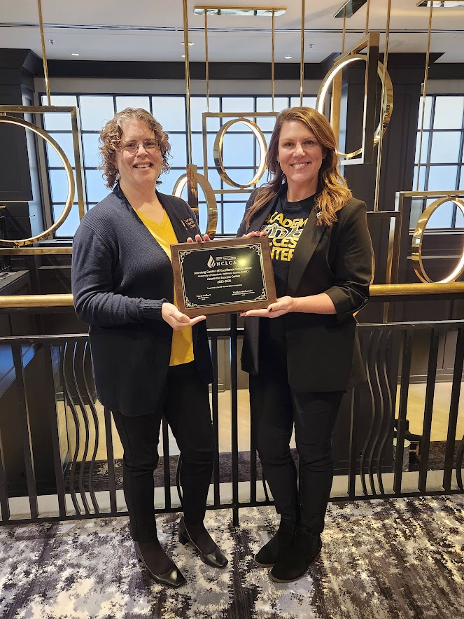 two women pose, holding a plaque between them