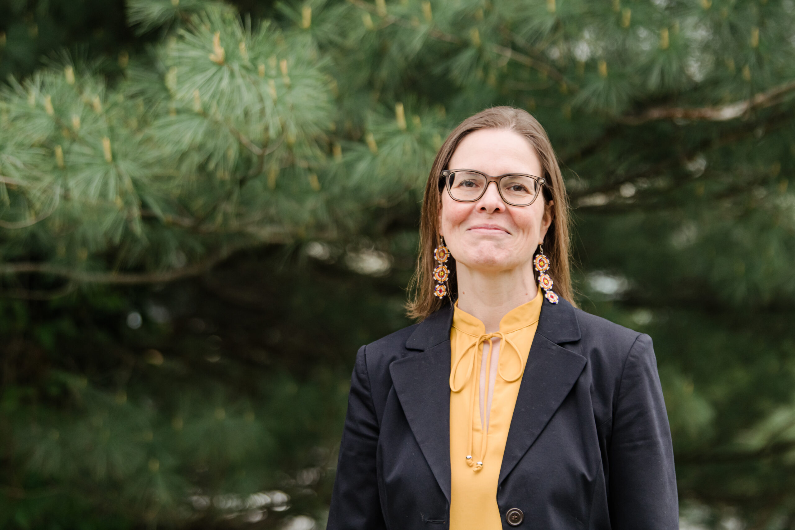 An adult wearing a black blazer and gold blouse stands outside in front of pine trees Academic Minute