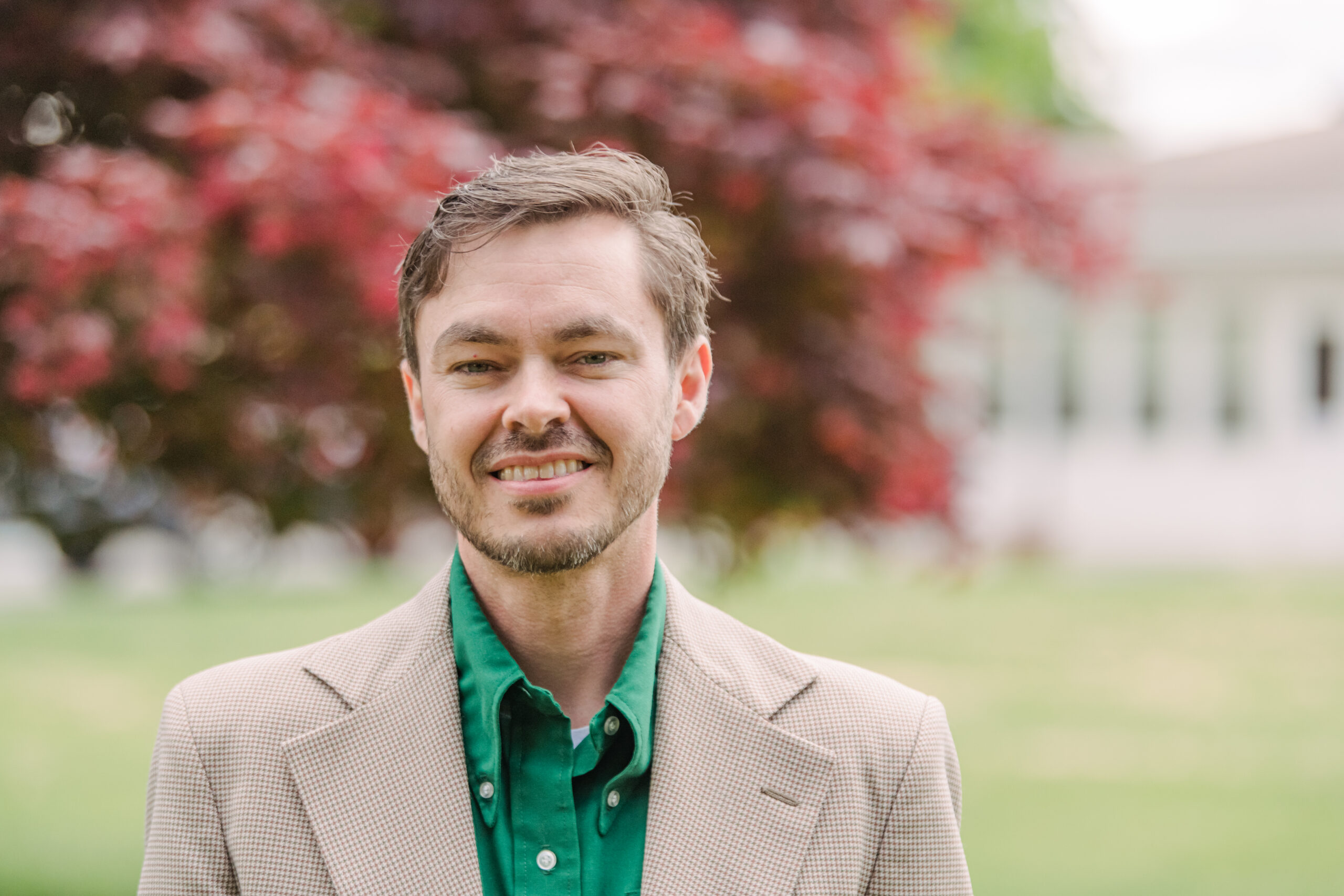 An adult with short brown hear wearing an emerald dress shirt and grey suit jacket stands outside in front of large pine trees. Academic Minute