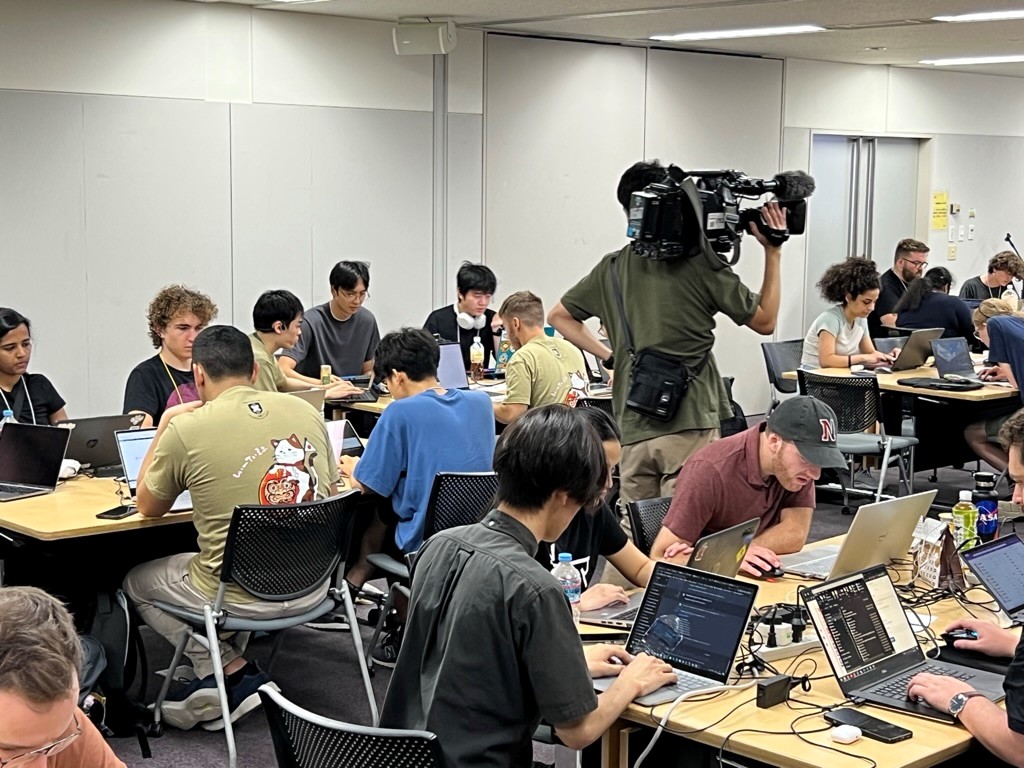 Many students sitting at desk with laptops in front of them competing during the 2023 Country-to-Country Capture the Flag competition at Keio University. There is a camera operator in the room with the competitors holding a large black television camera.
