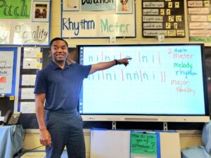 a teacher in a button up polo shirt instructs from the front of a music classroom