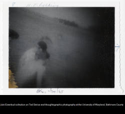 	
Thoughtograph. Faded image of person in hat and another person sitting near what appears to be water on left of image. Black spot near left border may be another head. Possibly a bridge structure in distance at top left of image. 