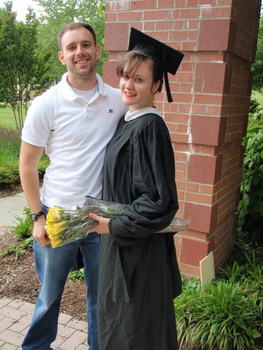 A man in a white polo shirt puts his arm around a woman in a grad cap and gown holding yellow flowers