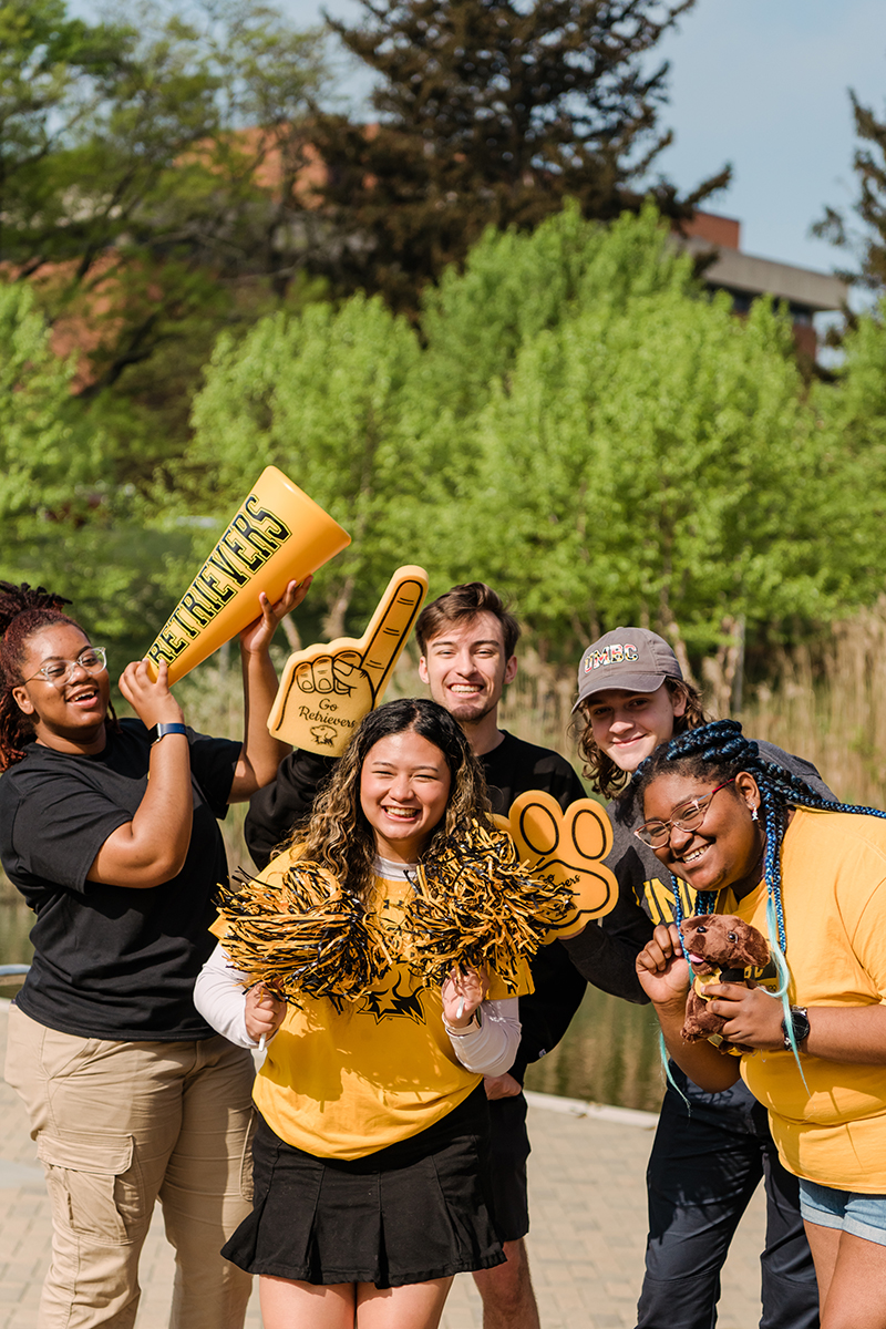 A diverse group of UMBC students, wearing black and gold spirit wear and posing with foam fingers and pom poms.