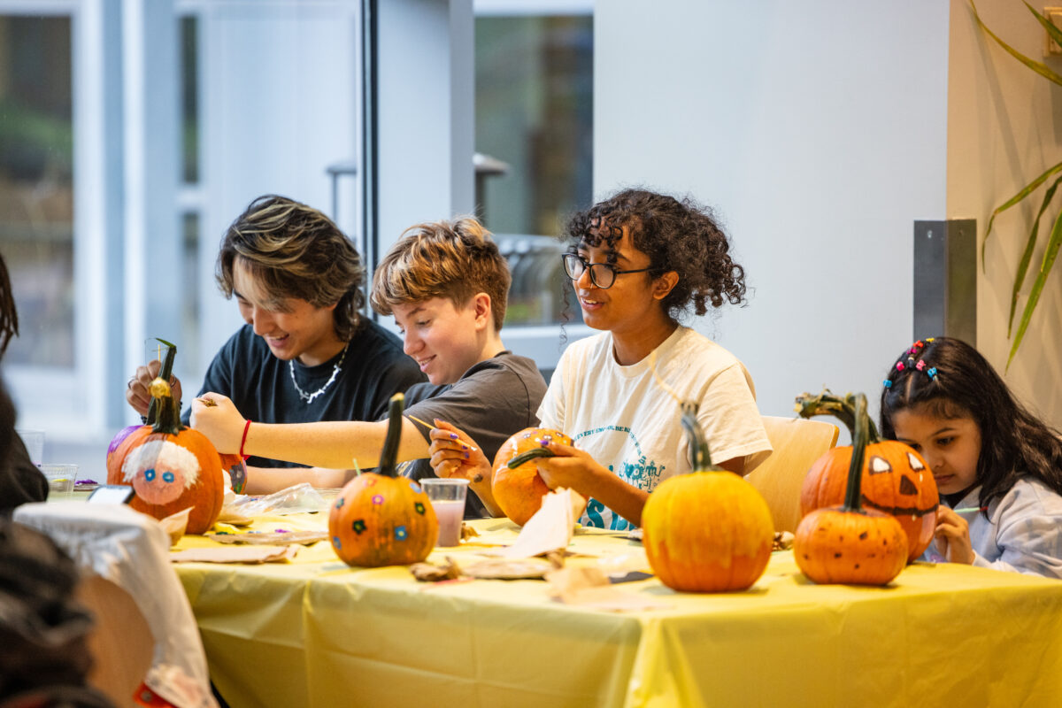 Children painting pumpkins indoors at a long table.