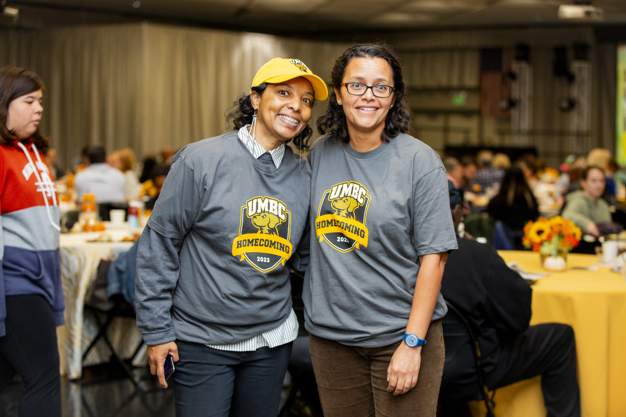 Two individuals stand smiling indoors wearing gray UMBC Homecoming t-shirts.