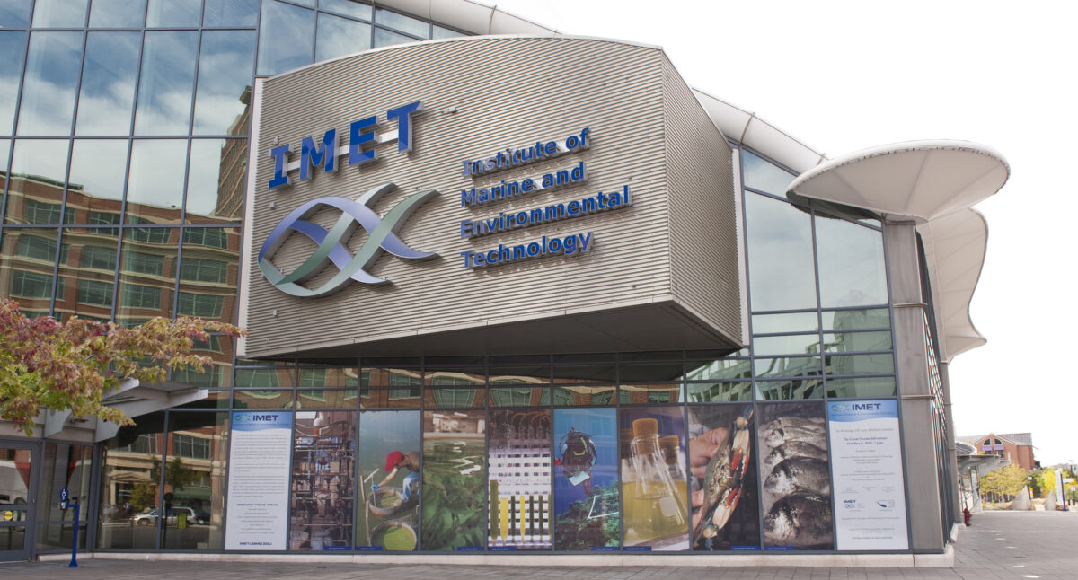 Building exterior; mostly glass with concrete protrusion labeled in blue with "IMET: Institute of Marine and Environmental Technology" and a logo of an abstract fish