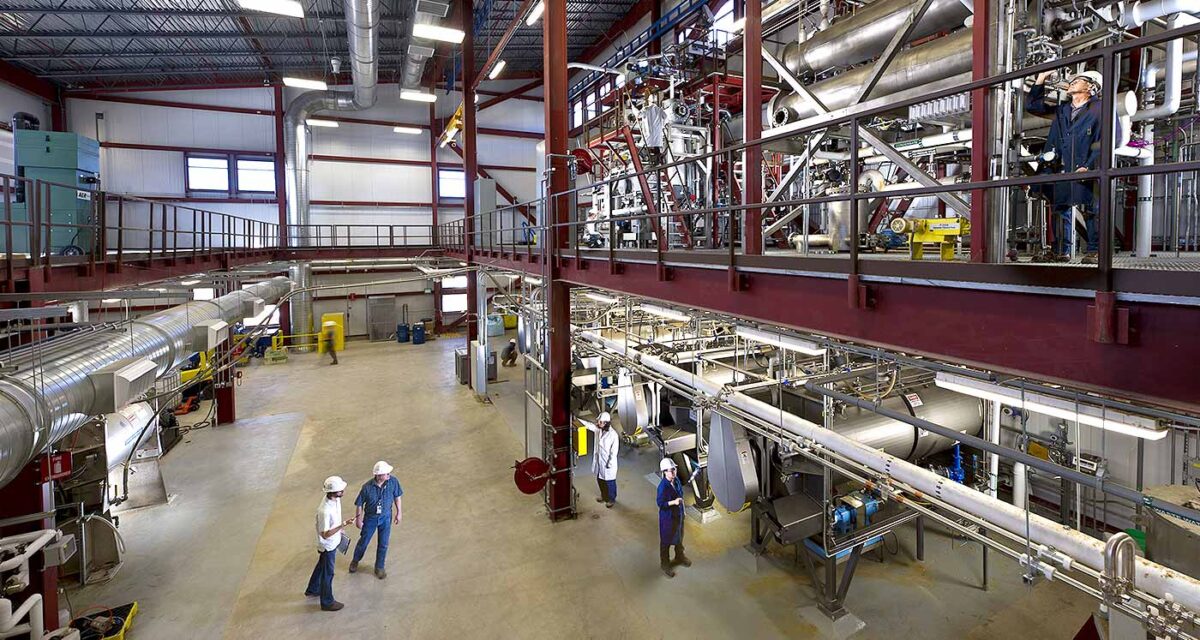 two-level large factory floor, many large shiny silver pipes, a few small people stand on the floor at the bottom