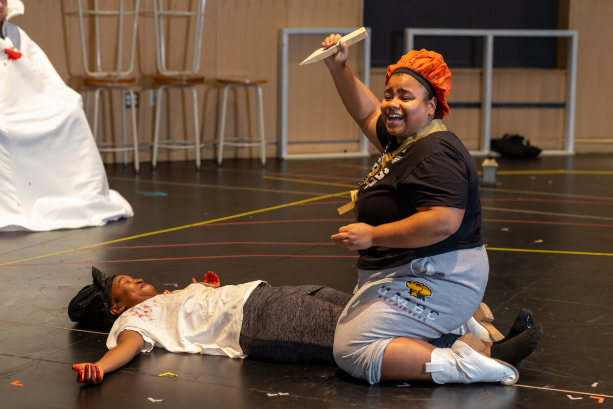 on a rehearsal stage, one actor straddles another actor holding a wooden stake in her hand