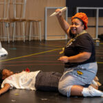 on a rehearsal stage, one actor straddles another actor holding a wooden stake in her hand