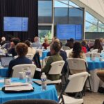 Open, naturally lit atrium with round tables with ocean blue tablecloths filled with people. Screens and podium at the front of the room; someone is presenting.